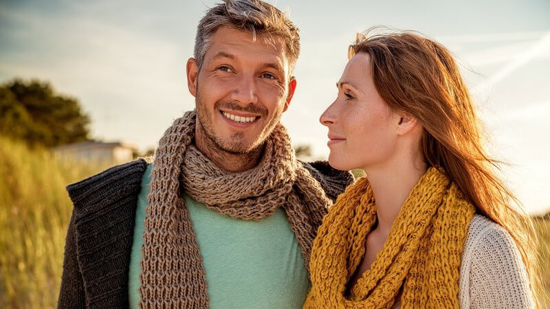 Paar im Feld mit Schals, sie schaut ihn glücklich an, er lächelt und sieht in die Ferne