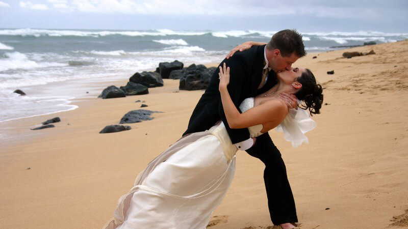 Frau im Brautkleid liegt Mann im schwarzen Anzug in den Armen, küssen sich leidenschaftlich am Strand