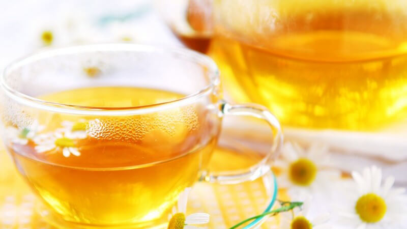 Glas und Kanne mit Kamillentee umgeben von Kamillenblüten