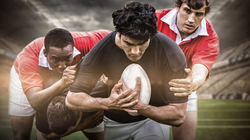 Ballführender Rugbyspieler in dunklem Trikot wird im Stadion von zwei Spielern in roten Trikots verfolgt