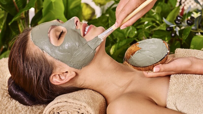 Frau liegt auf brauner Handtuchrolle und bekommt eine Tonerdemaske auf das Gesicht gepinselt