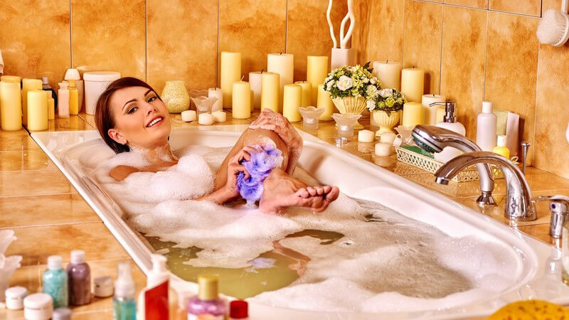 Frau liegt in Badewanne und wäscht ihre Beine mit einem Schwamm, umgeben von unzähligen Kerzen am Wannenrand