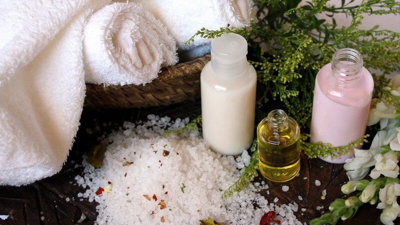 Badesalze, Öle und Cremes vor Kräutern und aufgerollten, weißen Handtüchern im Korb