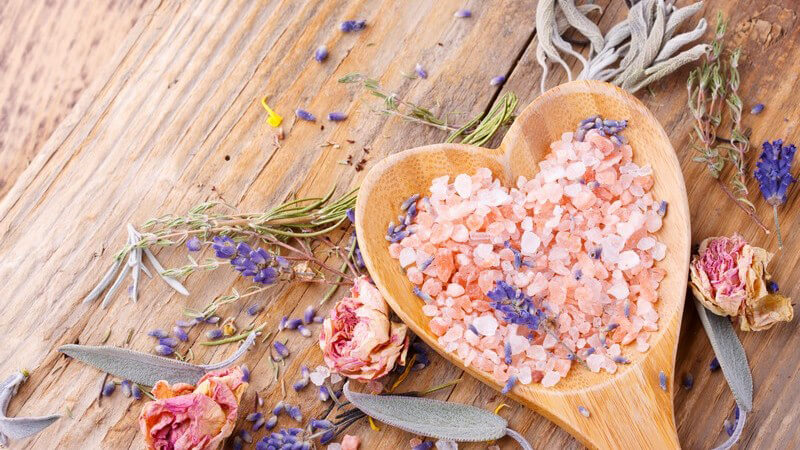 Badesalz auf herzförmigem Holzlöffel neben getrockneten Blüten und Kräutern