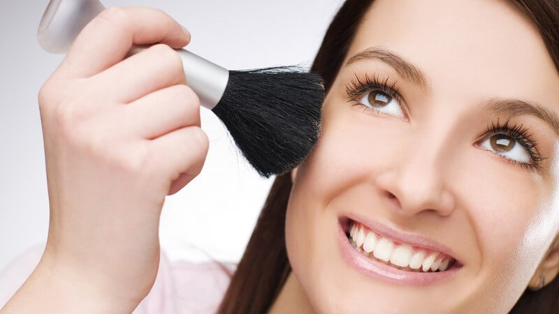 Junge Frau trägt Rouge mit Pinsel auf, lacht und schaut nach oben