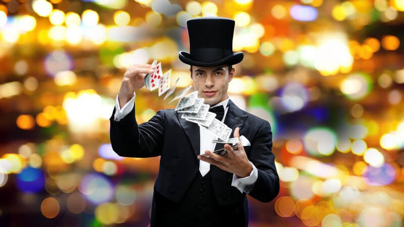 Zauberer in schwarzem Frack und Zylinder macht einen Trick mit Spielkarten, funkelnder Hintergrund