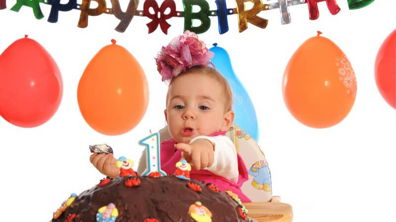 Baby am Geburtstag mit Kuchen, im Hintergrund Luftballons und "Happy Birthday"