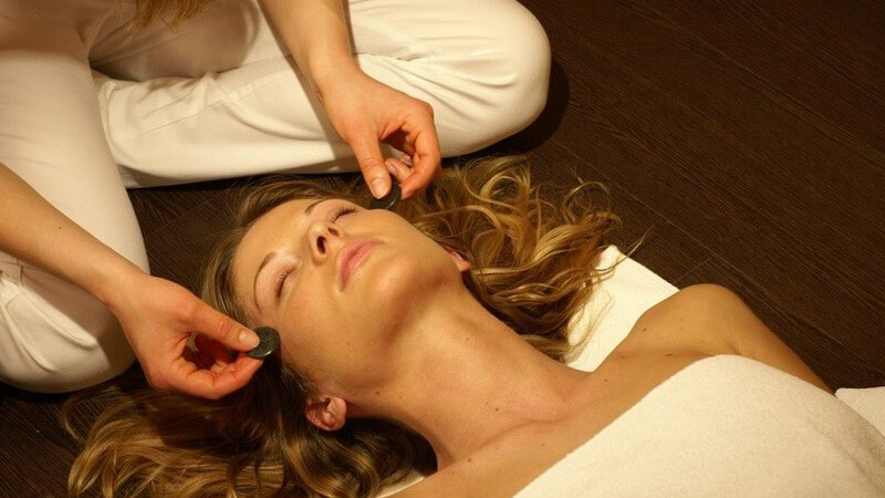 Frau mit geschlossenen Augen liegt auf Holzboden, in Handtuch, ihre Schläfen werden von zwei Händen mit Steinen massie