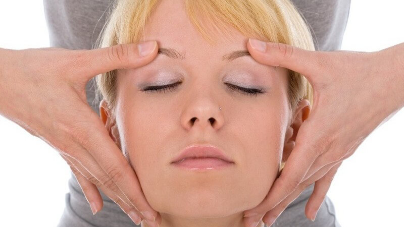 Frauengesicht einer blonden Frau, die eine dahinter stehenden Frau mit Händen an Schläfen massiert (Kopfmassage)