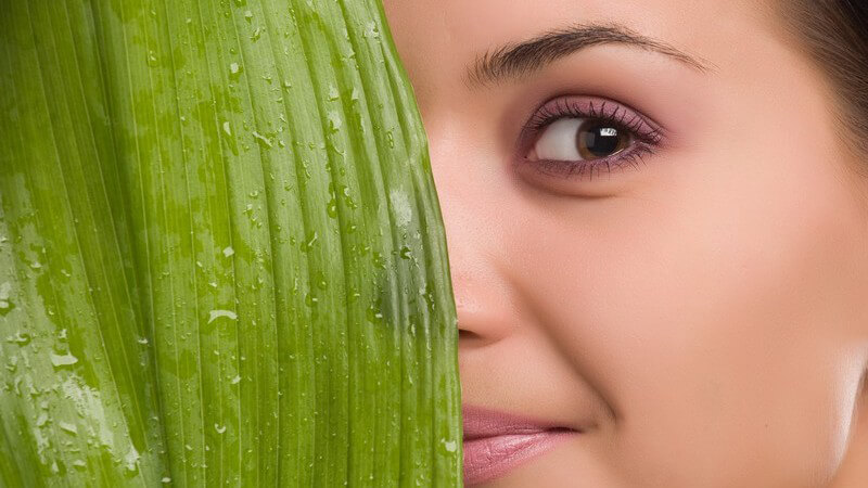 Gesicht einer dunkelhaarigen Frau versteckt sich halb hinter großem grünen Blatt, davor Wasser