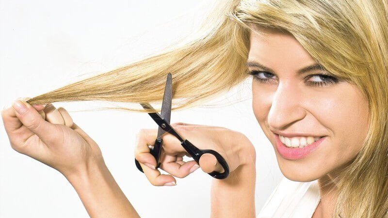 Blonde lächelnde Frau mit langen Haaren, die eine Schere ans Haar hält vor weißem Hintergrund