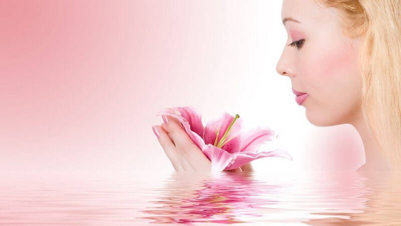 Linke Gesichtshälfte einer blonden jungen Frau, sieht auf rosa Lilie in ihren Händen, rosa Hintergrund in rosa Wasser