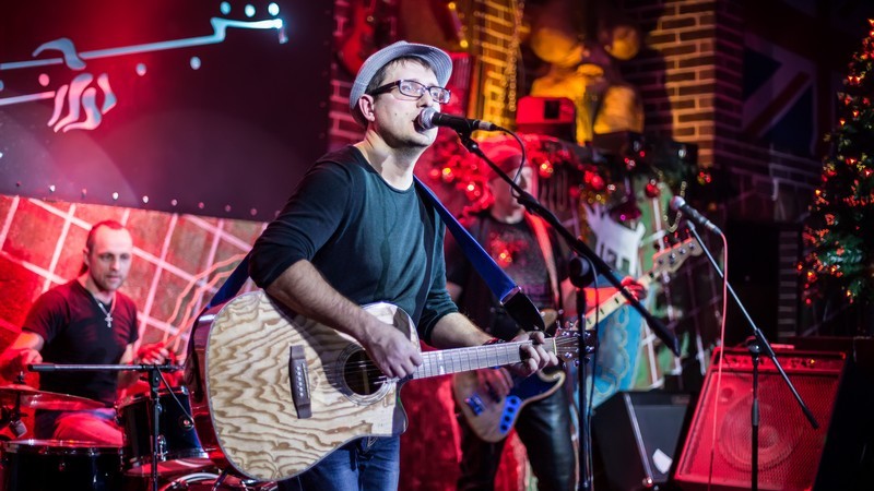 Musiker mit Brille und Hut singt und spielt Gitarre, dahinter ein Schlagzeuger und weiterer Gitarrist