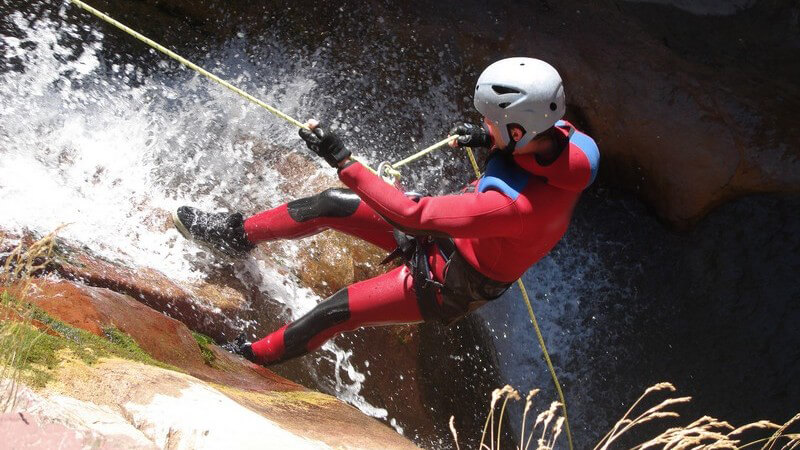 Person in rotem Neoprenanzug mit grauem Helm klettert gesichert an Felsen durch Wasserfall hoch