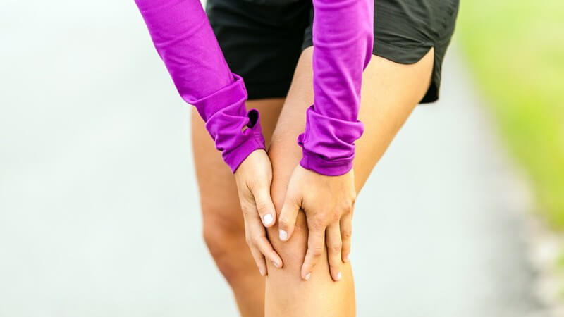 Joggerin in lila Laufbekleidung steht und fasst sich an das Knie