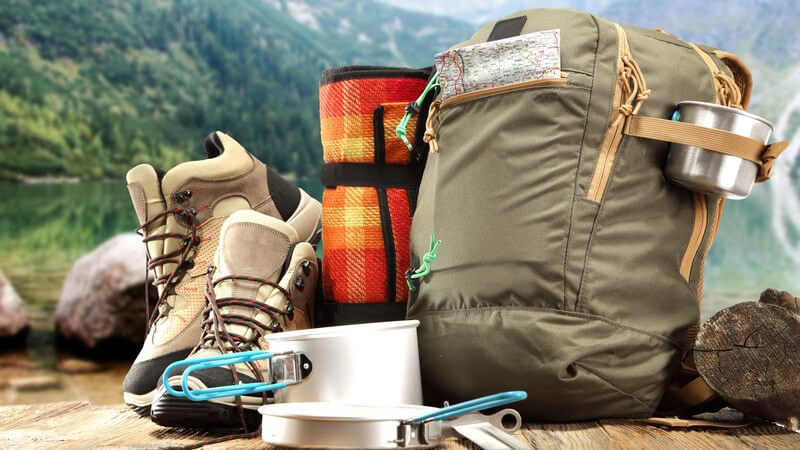 Wanderrucksack, Wanderschuhe und Campinggeschirr auf Holztisch vor Bergkulisse