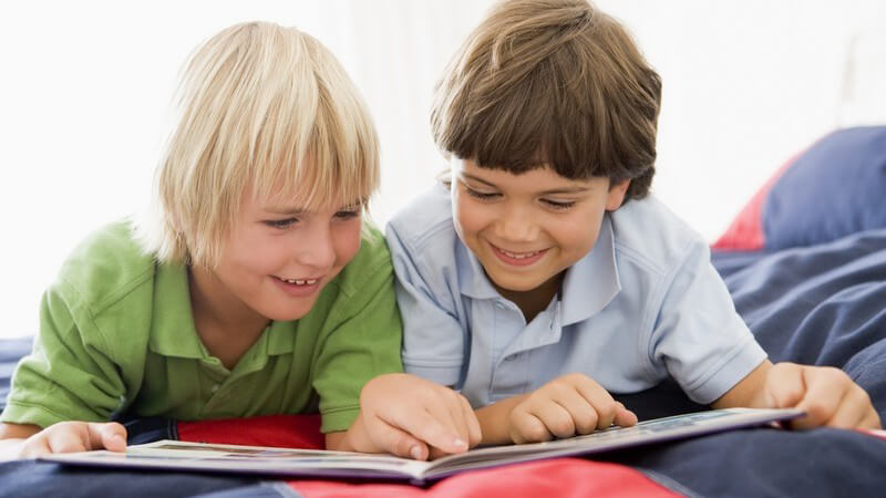 Zwei befreundete Jungen liegen lächelnd auf Bett und schauen sich gemeinsam ein Buch an
