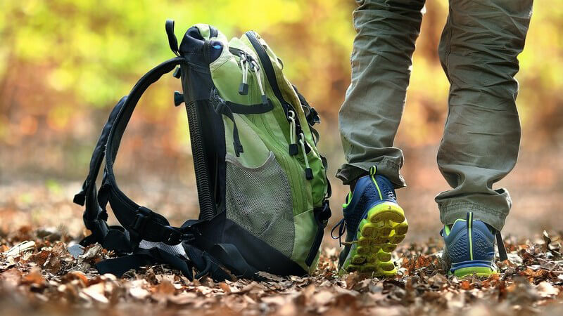 Beine mit blau-gelben Turnschuhen neben abgestelltem grünen Rucksack auf Waldweg mit viel Laub