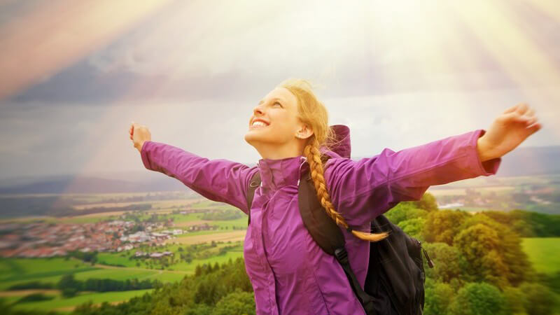 Frau mit Rucksack und lila Regenjacke steht auf einem Berg und blickt mit ausgebreiteten Armen zur Sonne