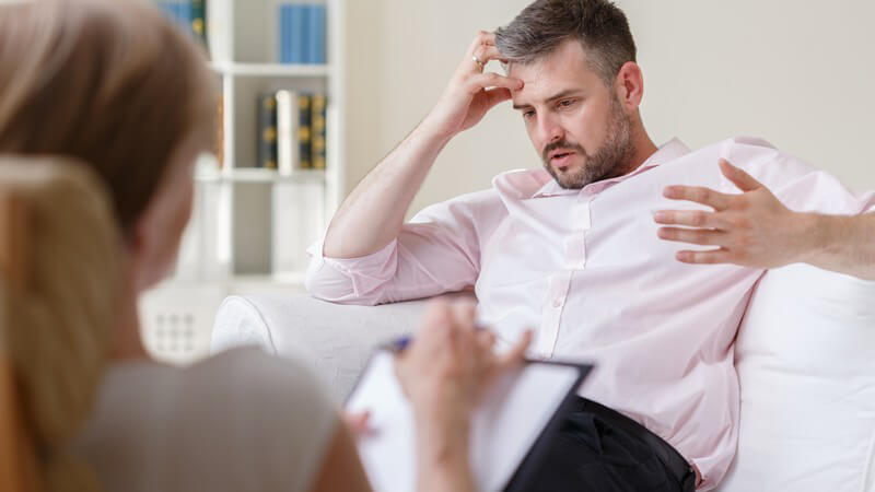 Geschäftsmann in rosa Hemd sitzt bei einer Psychotherapie auf der Couch, gegenüber die Psychotherapeutin mit Klemmbret