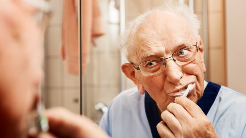 Alter Mann mit Brille steht im Badezimmer vor dem Spiegel und putzt sich die Zähne