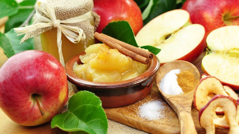 Kleines Schälchen mit Apfelmus und Zimtstangen, umgeben von frischen Äpfeln