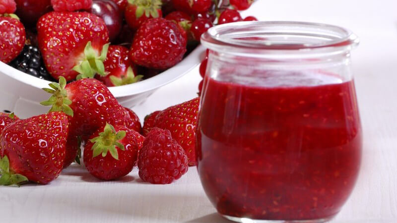 Glas mit Beeren Marmelade, dahinter Schale mit Früchten wie Erdberren, Himbeere etc.