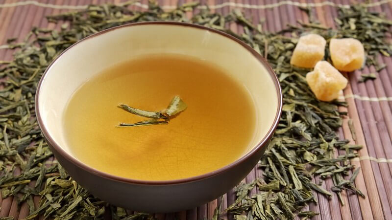 Schale mit grünem Tee auf Bastmatte mit losem Tee und Kandiszucker