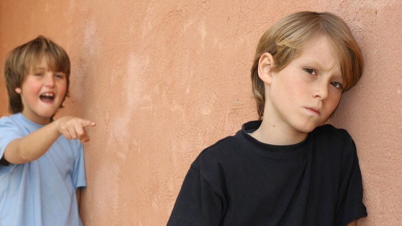 Trauriger Junge lehnt an Wand, hinter ihm zeigt Junge mit Finger auf ihn und lacht Ärgern Hänseln Mobbing Spott