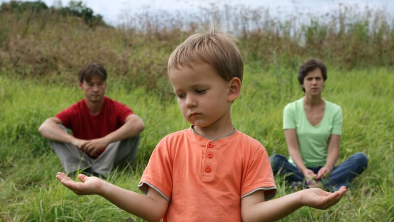 Trauriger Junge, die Hände zur Seite und Handflächen oben, im Hintergrund die Eltern